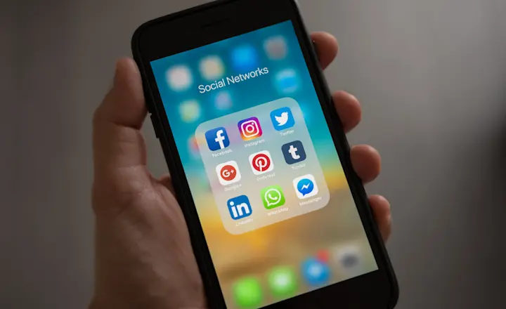 A hand holding a smartphone displaying a folder labeled "Social Networks." The screen shows various social media app icons, including Facebook, Instagram, Twitter, Google+, Pinterest, Tumblr, LinkedIn, WhatsApp, and Messenger.