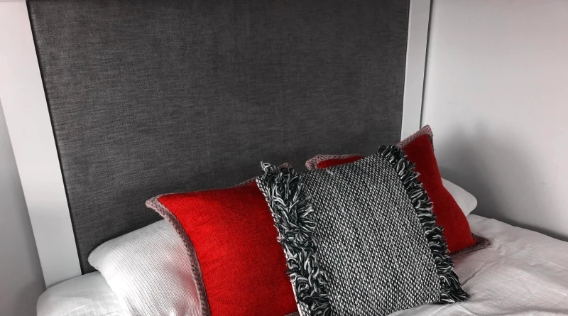 A bed with a gray fabric headboard features two red pillows with matching gray and red fringed edges, and a black and white woven decorative cushion in the center. The bedding is white, creating a modern and cozy look.