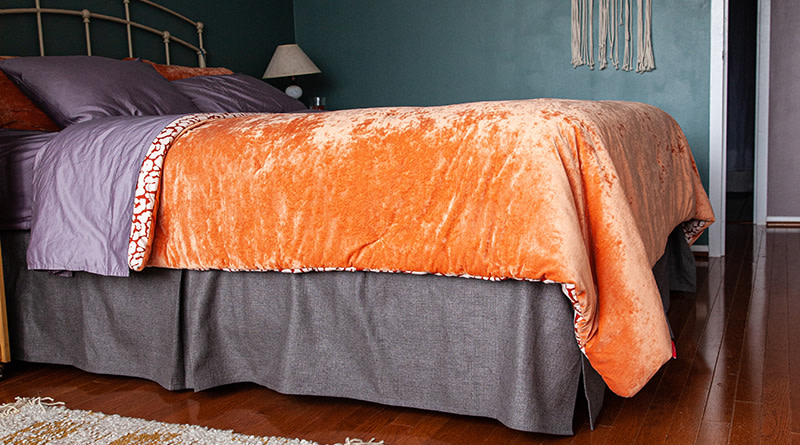 A grey bed skirt on a queen size bed with an orange comforter and light purple sheets in an apartment bedroom with blue/green walls.