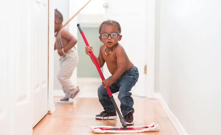 Two young children are cleaning a hallway. The child in the foreground wears jeans, glasses, and holds a mop. The child in the background is sweeping with a broom. The scene is indoors with wooden flooring and white walls.