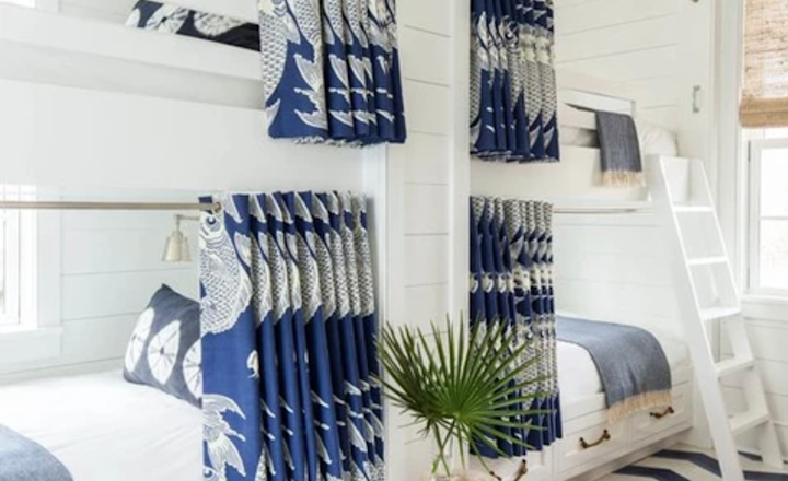 A cozy bedroom with white wooden walls features two bunk beds, each adorned with blue and white patterned curtains. A small tree trunk serves as a side table beside a leafy plant. The floor has a bold blue and white zigzag pattern.