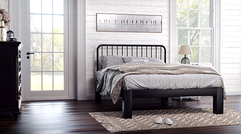 A black full size metal Farmhouse Bed in a quaint, rustic country home bedroom. Seen from the lower left-hand corner at a slight angle. Off-center in the image to the right.