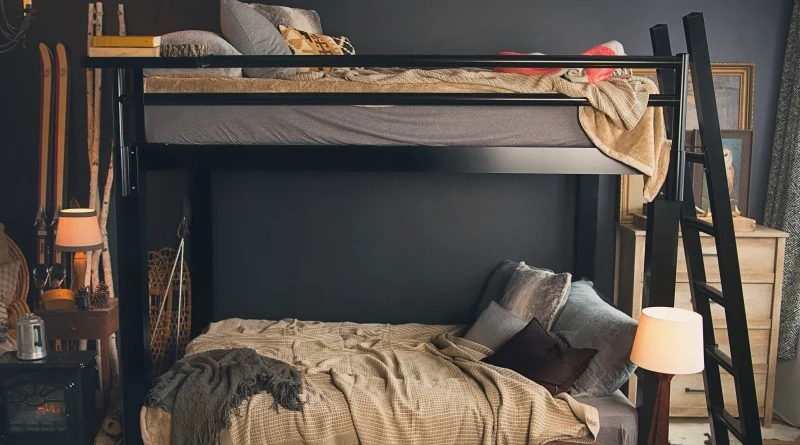 A cozy bedroom featuring a black metal loft bed with gray bedding on top and a double bed below with beige and brown pillows. The room includes wooden skis, a vintage snowshoe, a warm lamp, and dark gray walls.