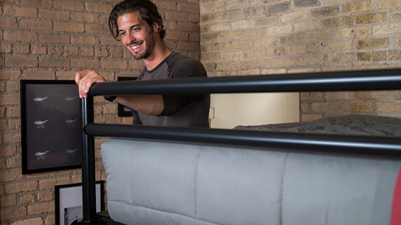 A man with shoulder-length hair smiles while leaning on the metal railing of a loft bed with a gray comforter. The room features brick walls and framed pictures in the background.