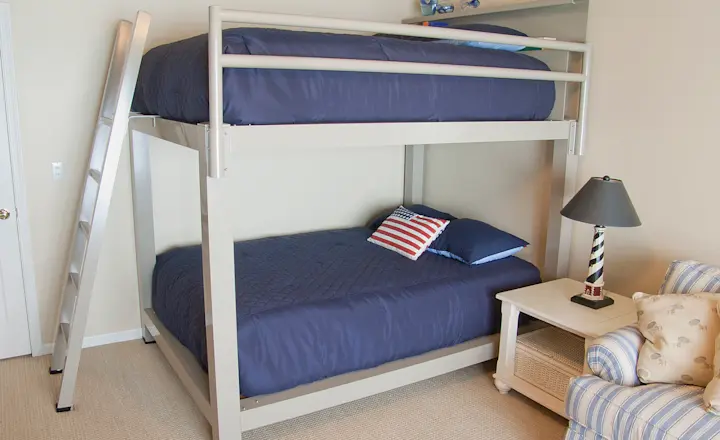 A bedroom with bunk beds featuring dark blue bedding and American flag-themed pillows. A ladder leads to the top bunk. The room includes a white nightstand with a striped lamp and a striped armchair with patterned pillows. Carpet covers the floor.