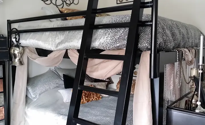 A bunk bed with silver and gold bedding, draped curtains, and decorative pillows. The top bunk is accessed by a black ladder. The room is adorned with metallic accents, candle holders, and decorative objects on a dresser beside the bed.