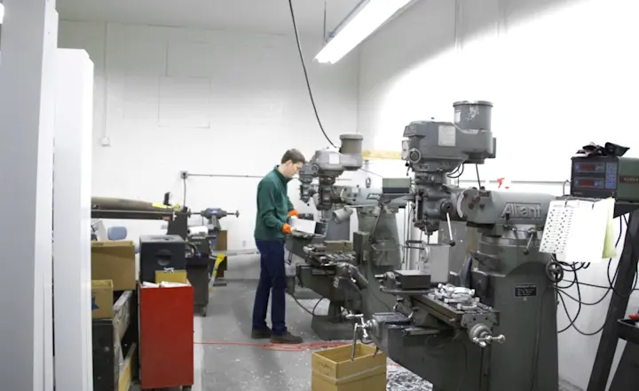 A person wearing safety gear operates a milling machine in an industrial workshop. Other machines and tools are visible in the neatly organized space, with bright fluorescent overhead lighting illuminating the area.