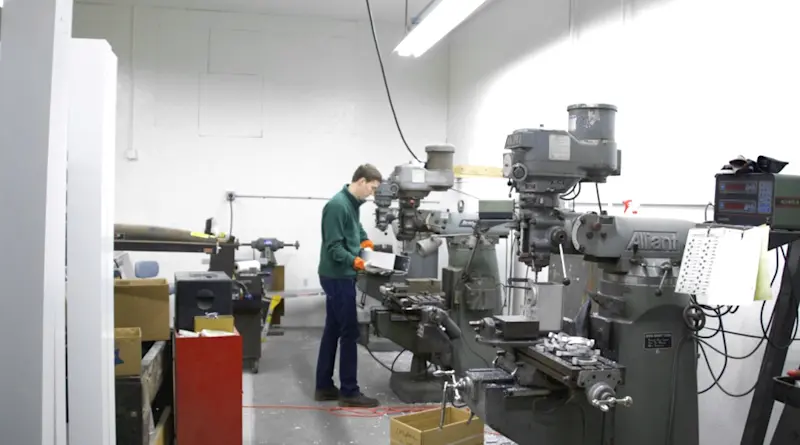 A person wearing safety gear operates a milling machine in an industrial workshop. Other machines and tools are visible in the neatly organized space, with bright fluorescent overhead lighting illuminating the area.