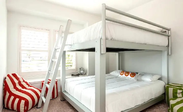 A small bedroom with a modern white bunk bed. The top bunk has a ladder while the bottom has decorative pillows. Two bean bag chairs, one red and white striped, sit in the corner near a window with shutters. The room has carpeted flooring.
