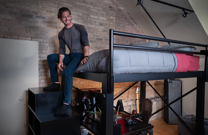 Young adult man sitting on the foot of his black Queen Loft Bed for adults resting his feet on the matching wooden staircase while he smiles and looks down toward the floor.