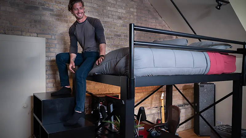 Young adult man sitting on the foot of his black Queen Loft Bed for adults resting his feet on the matching wooden staircase while he smiles and looks down toward the floor.