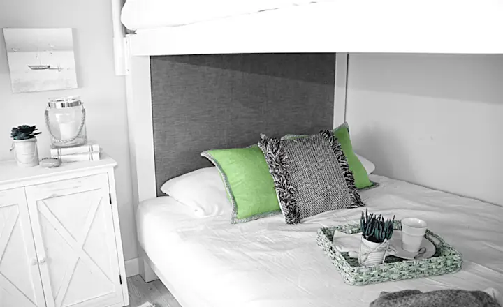 A cozy bedroom with a white bunk bed. The bottom bed has green and black accent pillows and a tray with a cup and decor. A white cabinet with a succulent and picture frame is beside the bed. The room has neutral and green tones.