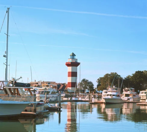 A marina with several boats docked, calm water reflecting the vessels and a red-and-white striped lighthouse in the background against a clear blue sky. Trees are visible near the lighthouse.