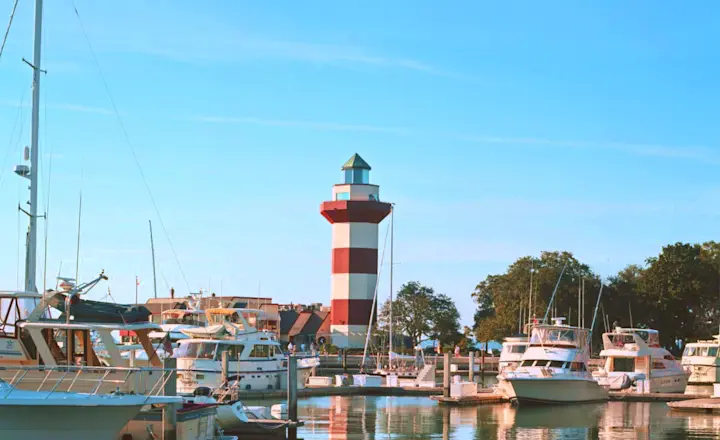 A marina with several boats docked, calm water reflecting the vessels and a red-and-white striped lighthouse in the background against a clear blue sky. Trees are visible near the lighthouse.