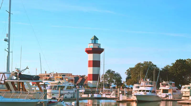 A marina with several boats docked, calm water reflecting the vessels and a red-and-white striped lighthouse in the background against a clear blue sky. Trees are visible near the lighthouse.