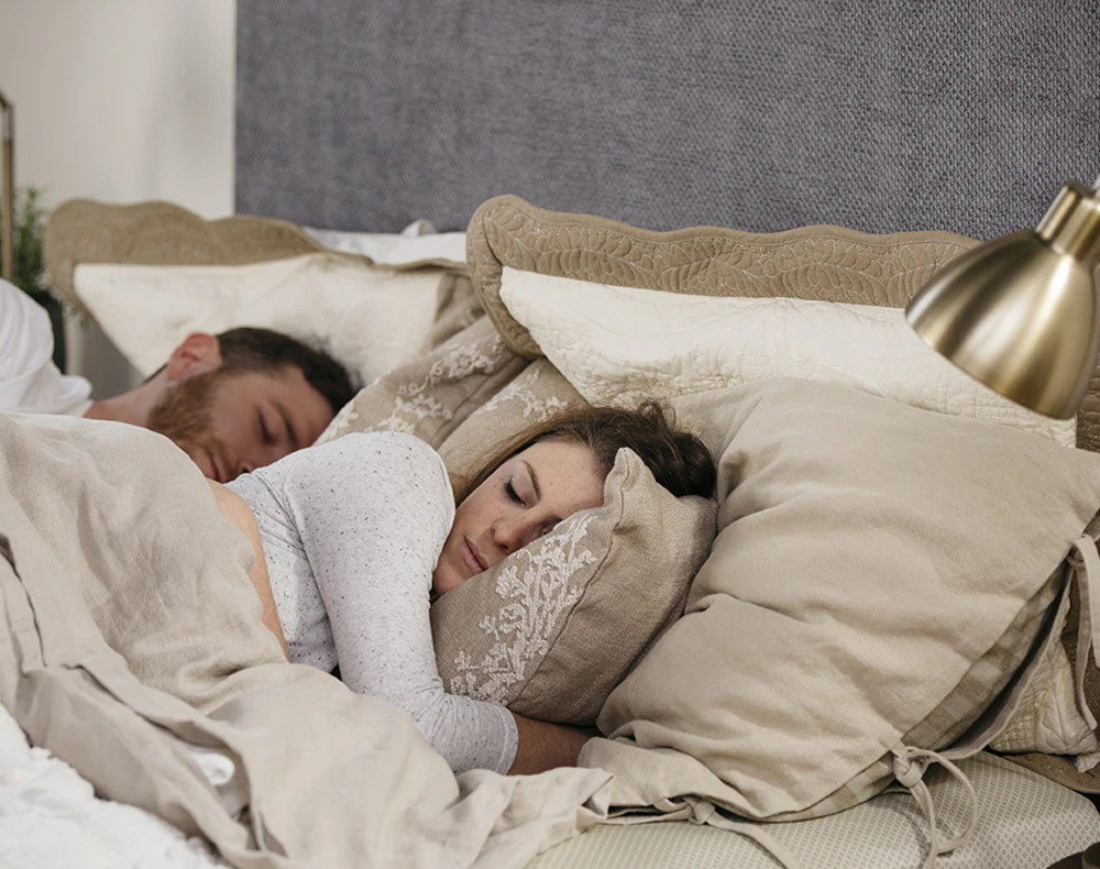 A young couple sleeping on their white queen size metal platform bed with a blueish gray headboard. Seen at a slight close-up from the right-hand side of the bed.