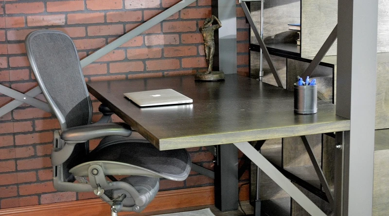 A modern, industrial-style workspace with a black desk against a brick wall. The desk has a closed laptop, a pen holder with blue pens, and a small sculpture. A gray ergonomic chair is in front of the desk.