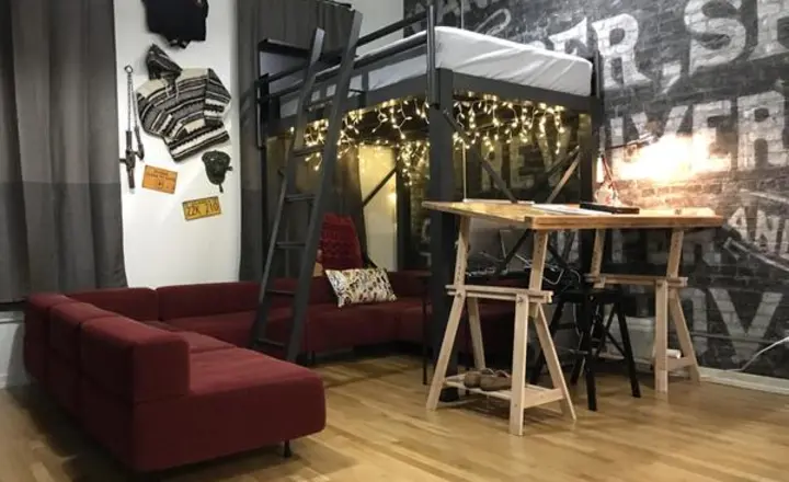 A cozy room with a loft bed decorated with string lights above a red sectional sofa. A wooden table with stools is nearby. Wall art and a textured accent wall add character. The room has hardwood floors and dark curtains.