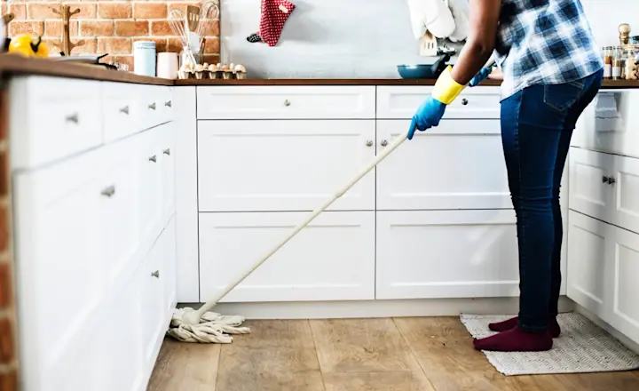 A person is mopping a kitchen floor. They are wearing gloves, blue jeans, and red socks. The kitchen has white cabinets and a brick backsplash. Cleaning supplies are seen on the countertop.