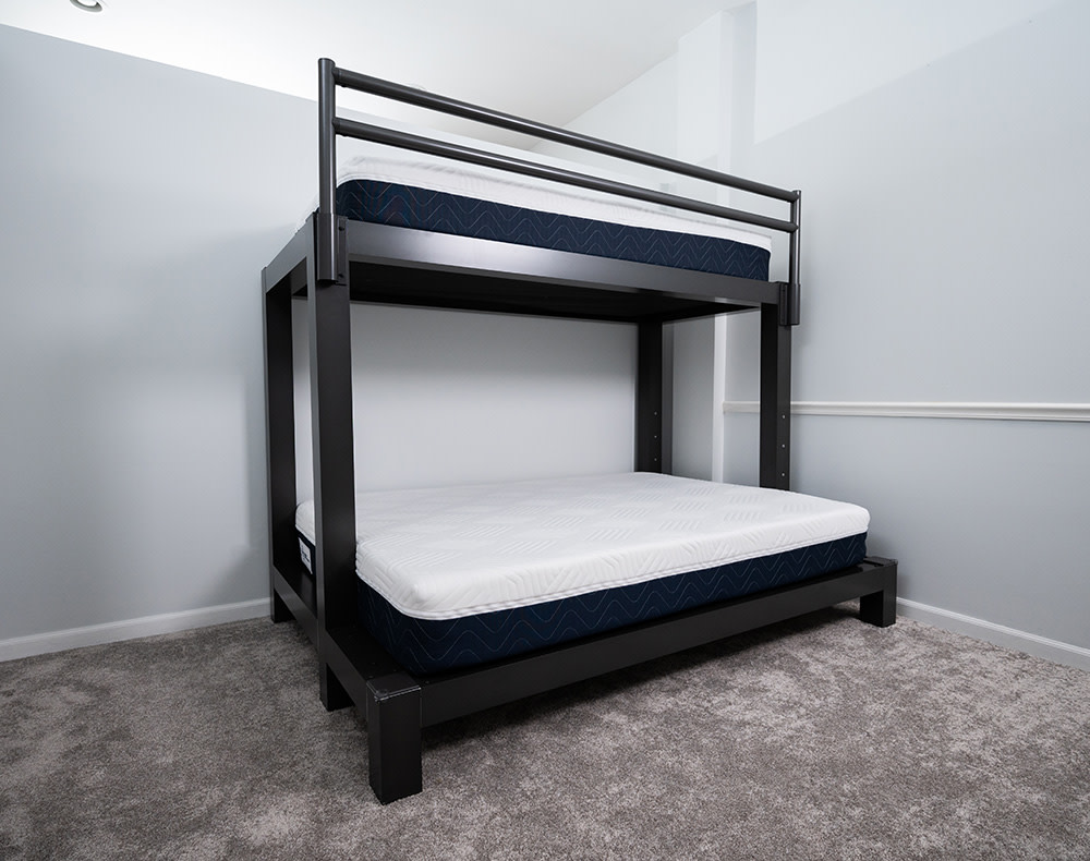 A charcoal colored Twin XL Over Queen Adult Bunk Bed in an empty carpeted room with light blue walls. Two uncovered Francis Lofts & Bunks mattresses rest on each bunk.