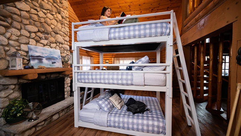 A cozy log cabin interior featuring a white triple bunk bed with blue and white gingham bedding. A person sits on the top bunk reading a book. Nearby, there is a stone fireplace with a painting and various decorations on the mantel.
