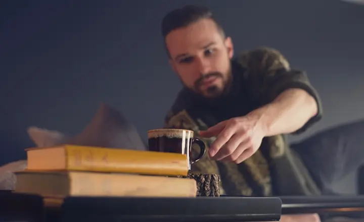 A person reaches for a frothy coffee mug resting on a stack of books, against a blurred background. The scene suggests a cozy and relaxed atmosphere.