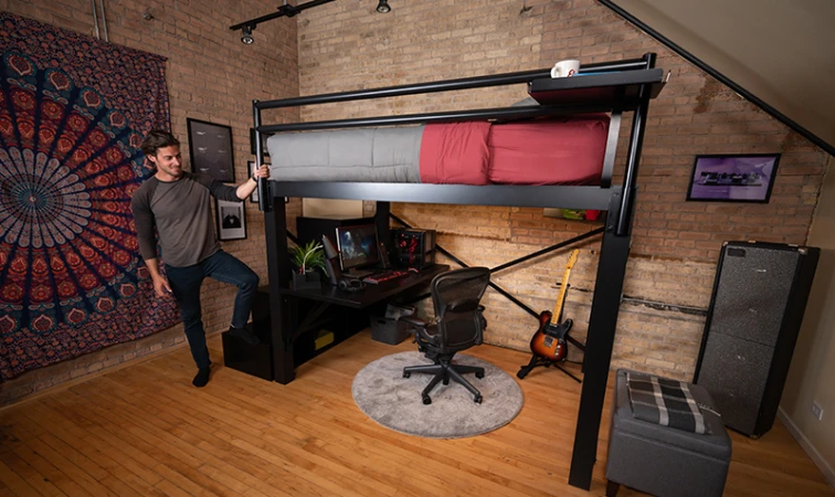 Man standing on the bottom stair of the staircase of a black Queen size Adult Loft Bed in his studio apartment and holding the guard rail as he looks at the space beneath the bed frame.