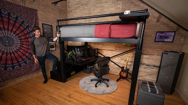 Man standing on the bottom stair of the staircase of a black Queen size Adult Loft Bed in his studio apartment and holding the guard rail as he looks at the space beneath the bed frame.
