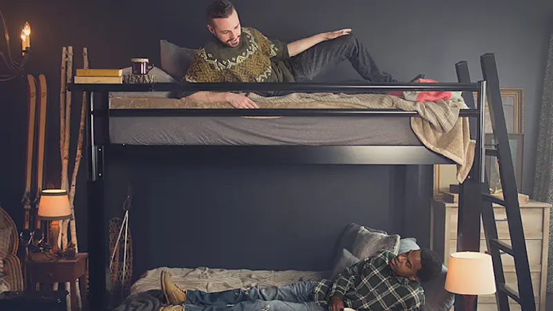 Two Young adult men on a black Queen Over Queen Bunk Bed for adults in a ski lodge bedroom, each lying on a bunk and looking at each other talking.
