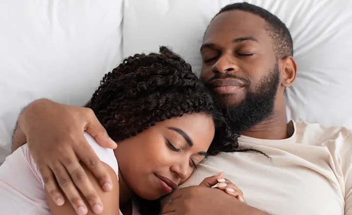A young black couple lying in bed together asleep on all-white bedding with the woman's head resting on the man's shoulder. His right arm is around her shoulders. They're seen from a high angle directly above their heads.