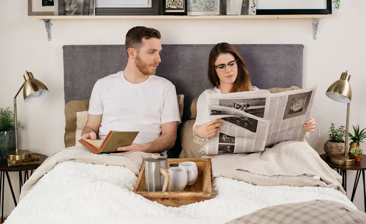 A man and a woman are sitting in bed under a light comforter. The man is holding a book, and the woman is reading a newspaper. A wooden tray with a coffee set and a plant is on the bed. Lamps and framed pictures are in the background.