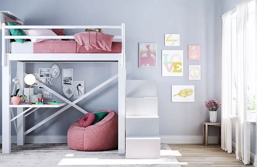 A bright room with a white loft bed featuring pink bedding. A workspace with a desk, chair, and decorations is underneath. The wall has colorful framed art, and sunlight streams in through a window with a pink bean bag and a small table nearby.