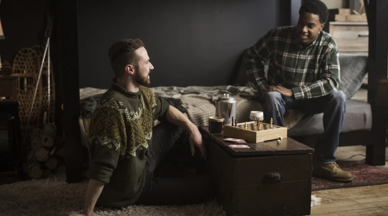 Two men sit talking, one on the bottom bunk of a black Queen Over Queen Adult Bunk Bed, and one on the floor looking up at the other.