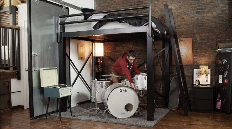 A young man sets up a white acoustic drum kit underneath his black Adult Loft Bed in his urban apartment.