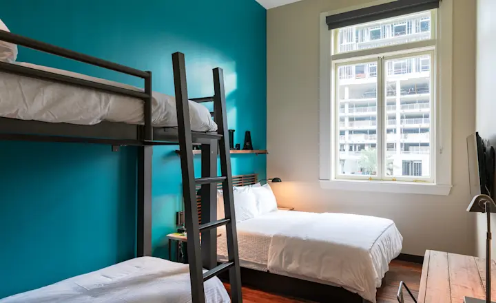 A cozy bedroom with teal and beige walls features a bunk bed and a double bed. Natural light streams through a large window, illuminating a wooden floor. The room includes a small lamp, a TV, and a wooden desk.