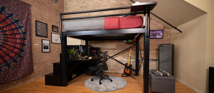 Black Queen Adult Loft Bed with a staircase and attached desk and red and gray bedding in a studio apartment with brick exposed walls seen from a lower angle at a slight distance from the upper left-hand side of the bed.
