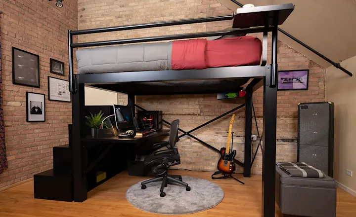 Black Queen Adult Loft Bed with a staircase and attached desk and red and gray bedding in a studio apartment with brick exposed walls seen from a lower angle at a slight distance from the upper left-hand side of the bed.