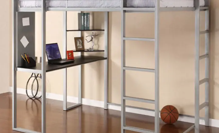A loft bed with a metal frame and a ladder. Underneath, there's a desk with a few books, a laptop, and decorative items. A basketball is on the wooden floor nearby. The walls are light-colored, with a few framed pictures hanging.
