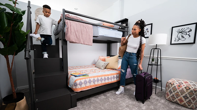 Young mother standing in the a nice guest bedroom next to a charcoal Adult Bunk Bed holding a suitcase and talking to her young son standing at the top of the wooden storage staircase holding the aluminum handrail.