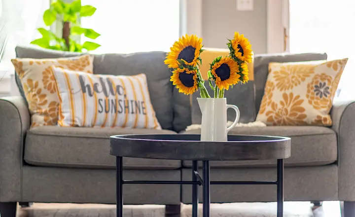 A cozy living room with a gray sofa adorned with yellow and white pillows, two of which read "hello sunshine." A round coffee table in front holds a vase of sunflowers. A tall plant stands to the left, and sunlight brightens the scene.