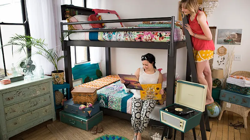 Two young adult women in a shared apartment bedroom on a charcoal Queen Over Queen Bunk Bed for adults with one of them sitting on the bottom bunk looking at vinyl records and the other standing on the ladder looking over her shoulder.