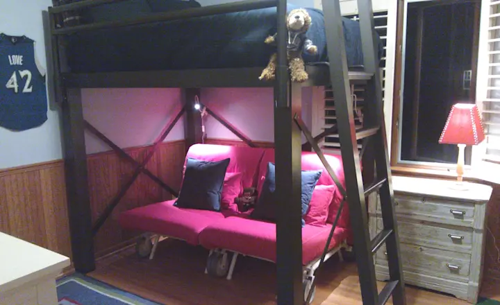 A bedroom with a black loft bed and a ladder. Below, there's a pink futon with blue and pink pillows. A stuffed animal sits on the bed. A wooden dresser with a red lamp is on the right, and sports jersey decorates the wall.
