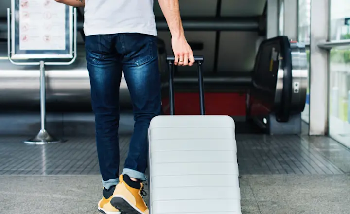 A person in jeans and a white T-shirt is walking inside an airport terminal, pulling a silver suitcase. An escalator and a sign are visible in the background.