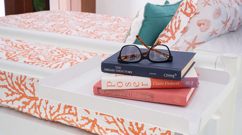 A white tray attached to a white metal bed frame with three books stacked on it.