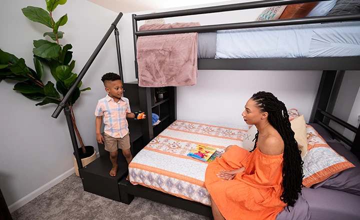 Charcoal Adult Bunk Bed in a simple guest room with a young mother sitting on the bottom bunk talking to her 4-year-old son as he stands on the bottom stair of the matching wooden staircase.