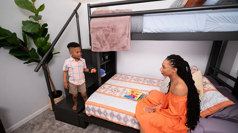 Charcoal Adult Bunk Bed in a simple guest room with a young mother sitting on the bottom bunk talking to her 4-year-old son as he stands on the bottom stair of the matching wooden staircase.