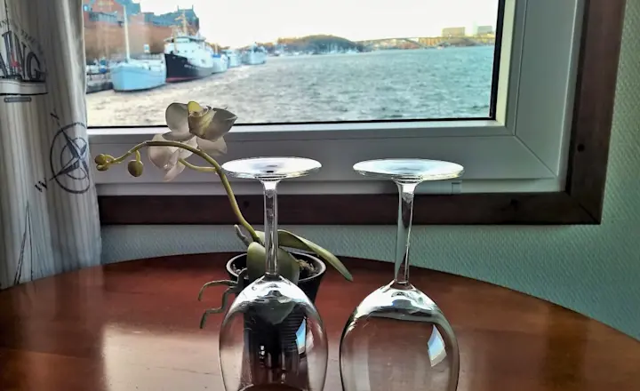 Two upside-down wine glasses sit on a wooden table next to a small potted orchid. A large window in the background offers a view of a river, boats, and buildings on a cloudy day.