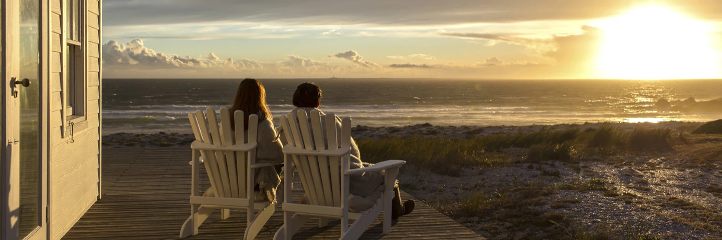 Two adults sitting facing away from the camera in white beach chairs on the large wooden desk of a beach house watching the sunset from across the beach.