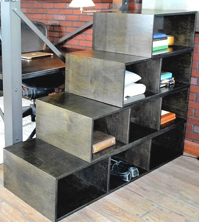 A wooden staircase shelf unit with storage compartments is filled with various items like books, a folded towel, and a pair of shoes. It is situated in a room with a brick wall and a desk in the background.