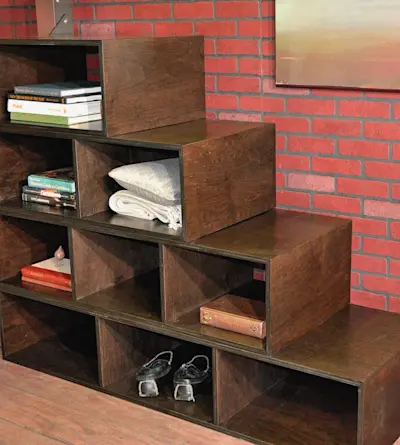 A dark wooden, staircase-style shelf against a red brick wall, holding books, a decorative pillow, a small sculpture, and shoes at the bottom. The shelf has an industrial, modern design.